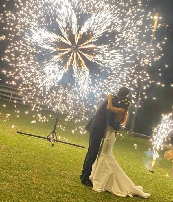 Coppia che si abbraccia sotto i fuochi d'artificio durante un matrimonio all'aperto. I fuochi d'artificio esplodono a forma di stella, illuminando la coppia su un prato erboso.