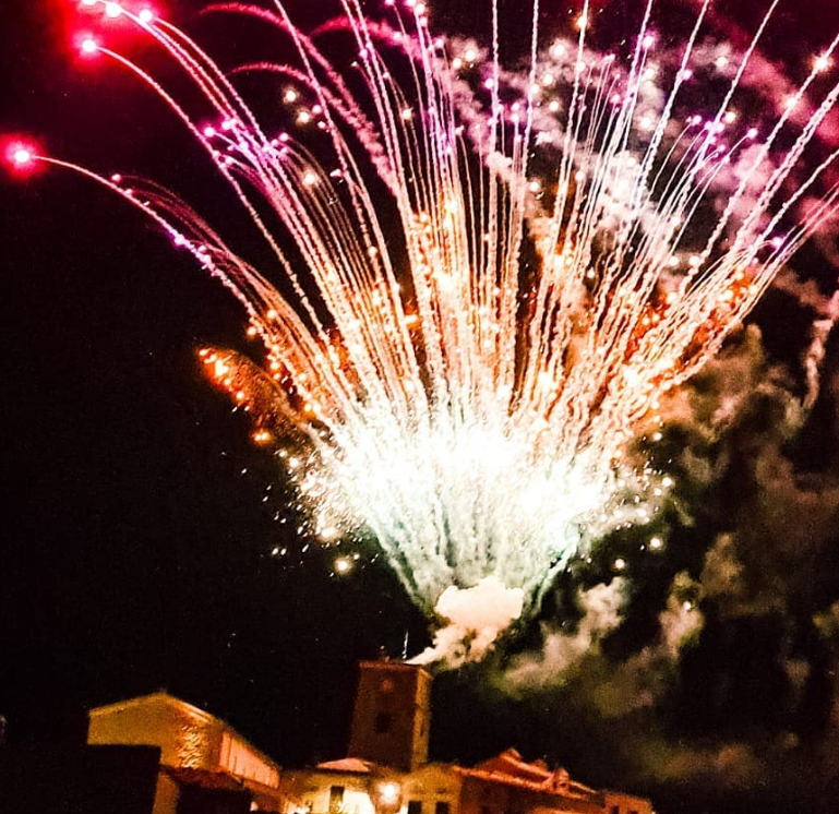 Di notte, i fuochi d'artificio esplodono sopra un edificio: esplosioni rosse, rosa, arancioni e bianche contro un cielo scuro.