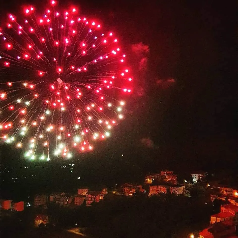 Spettacolo pirotecnico notturno sopra una città: esplosioni rosse e bianche illuminano il cielo.