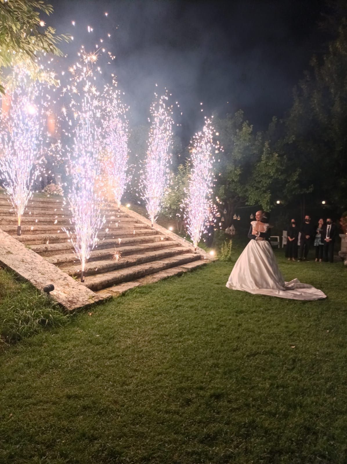 Una sposa in abito bianco osserva i fuochi d'artificio esplodere vicino ai gradini di pietra durante un matrimonio all'aperto.