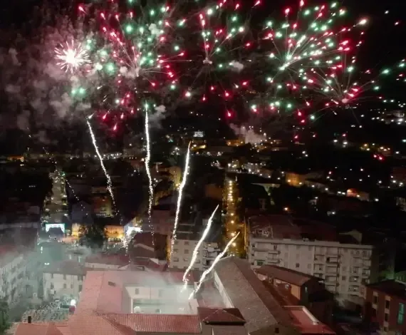 Fuochi d'artificio su una città di notte. Esplosioni rosse, verdi e bianche con scie su edifici e luci.