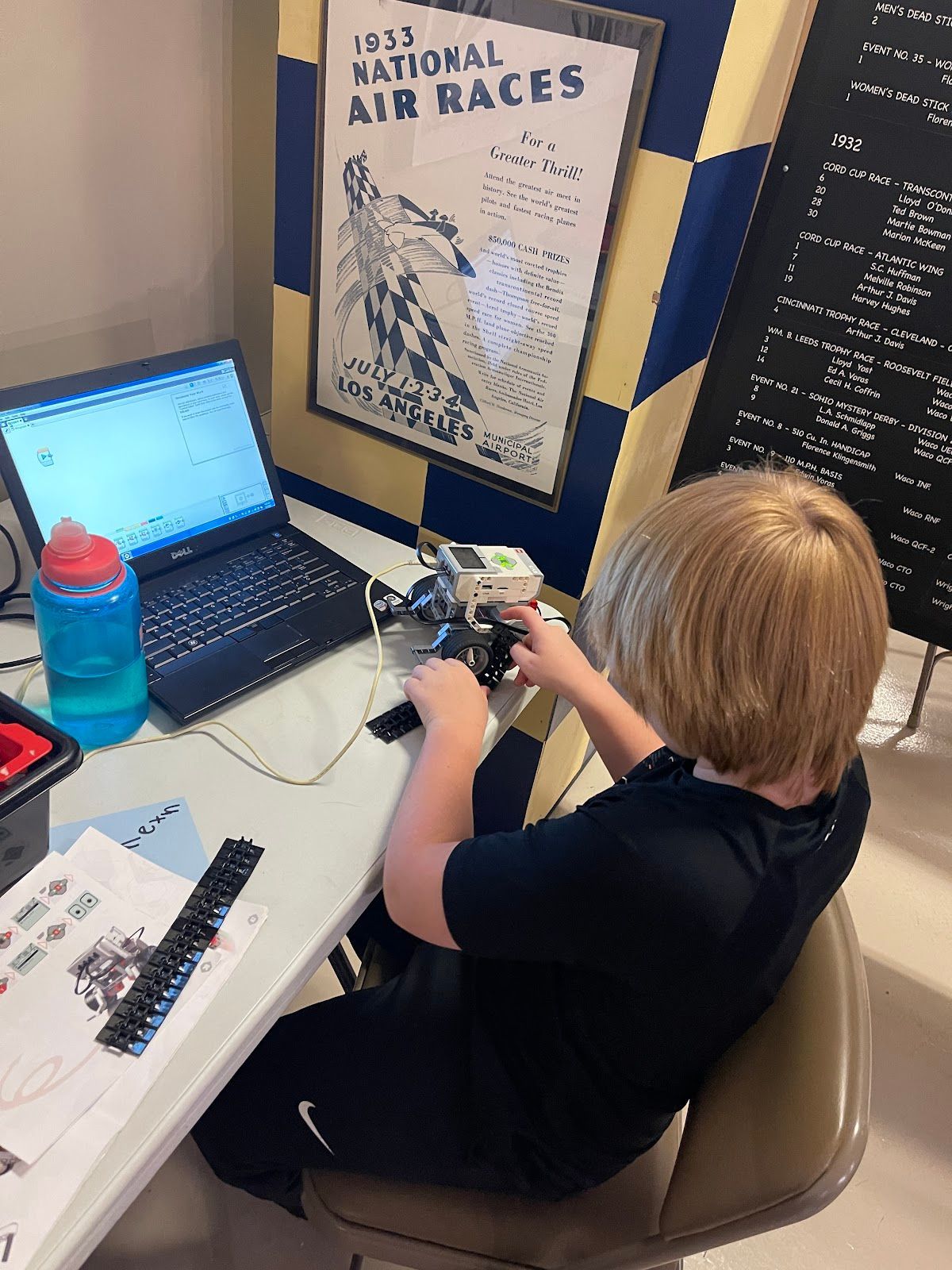 A young boy is sitting at a desk with a laptop and a poster on the wall that says 1933 national air races