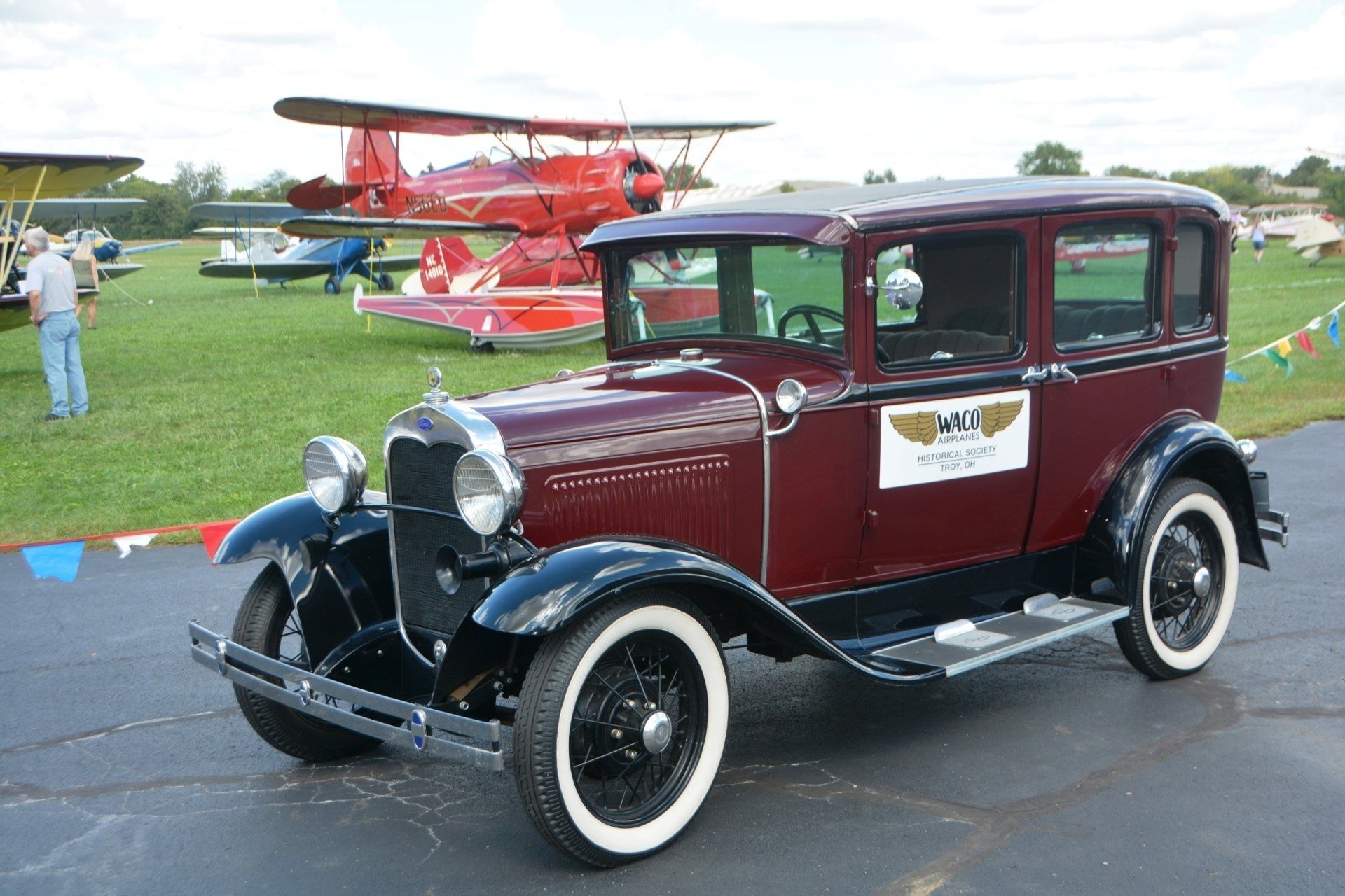 An old car is parked in front of a field of planes.