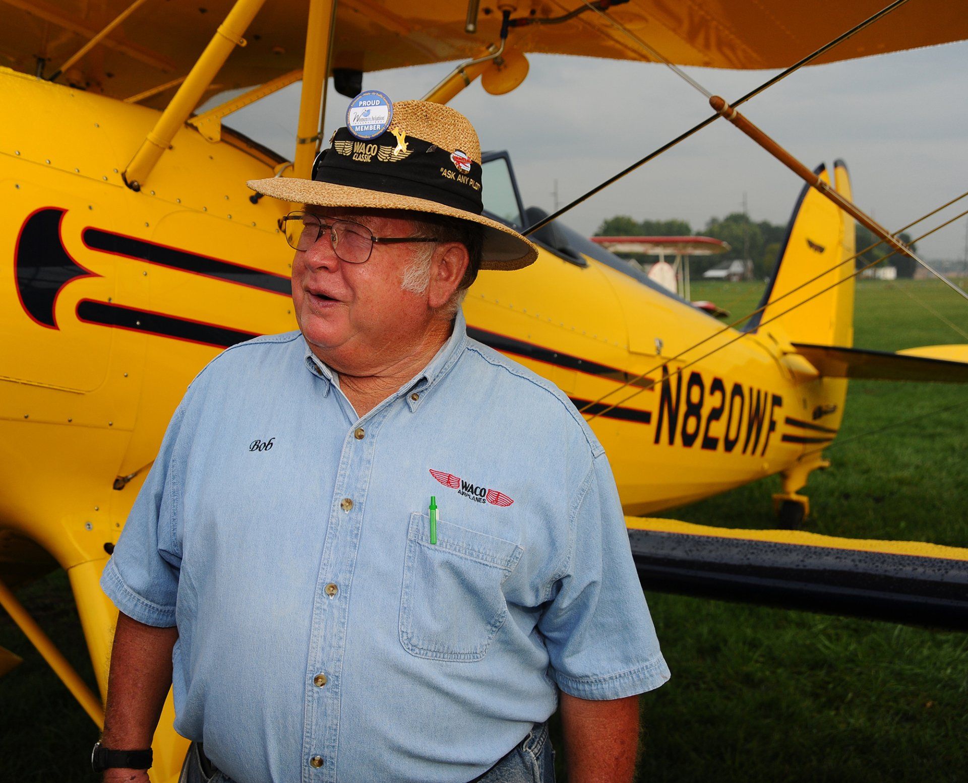 A man in a straw hat stands in front of a yellow plane with n820wf on the tail