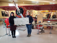 Two women are holding a large check in front of an airplane.