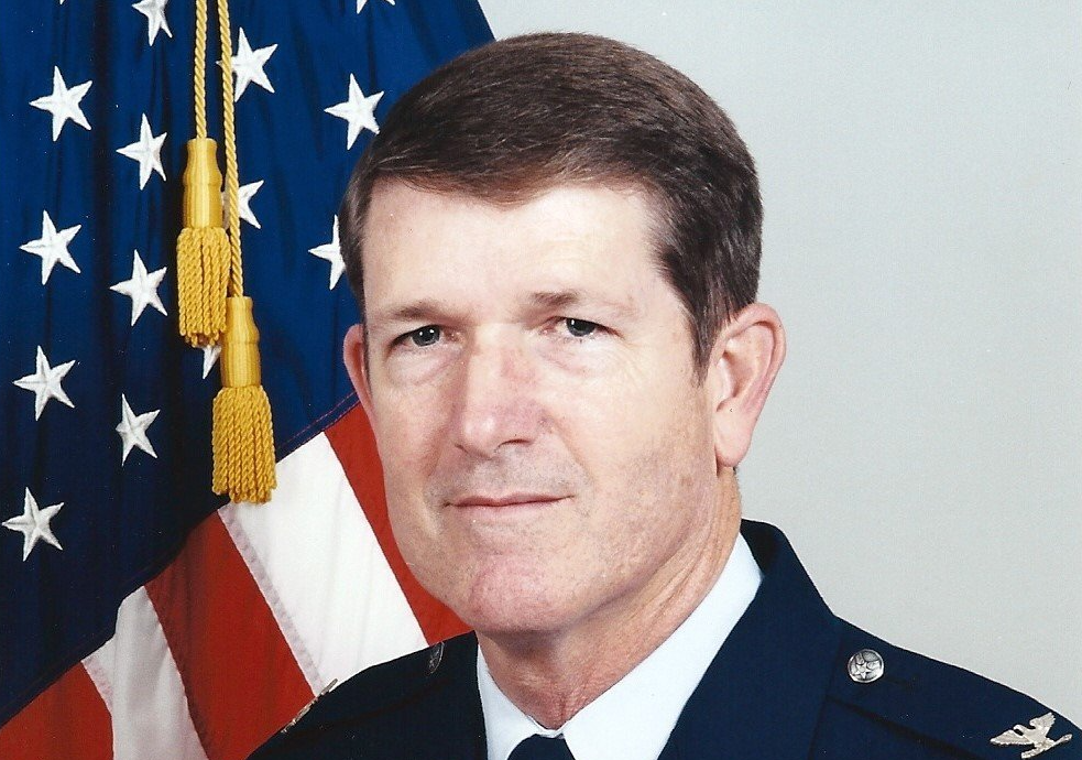 A man in a military uniform stands in front of an american flag