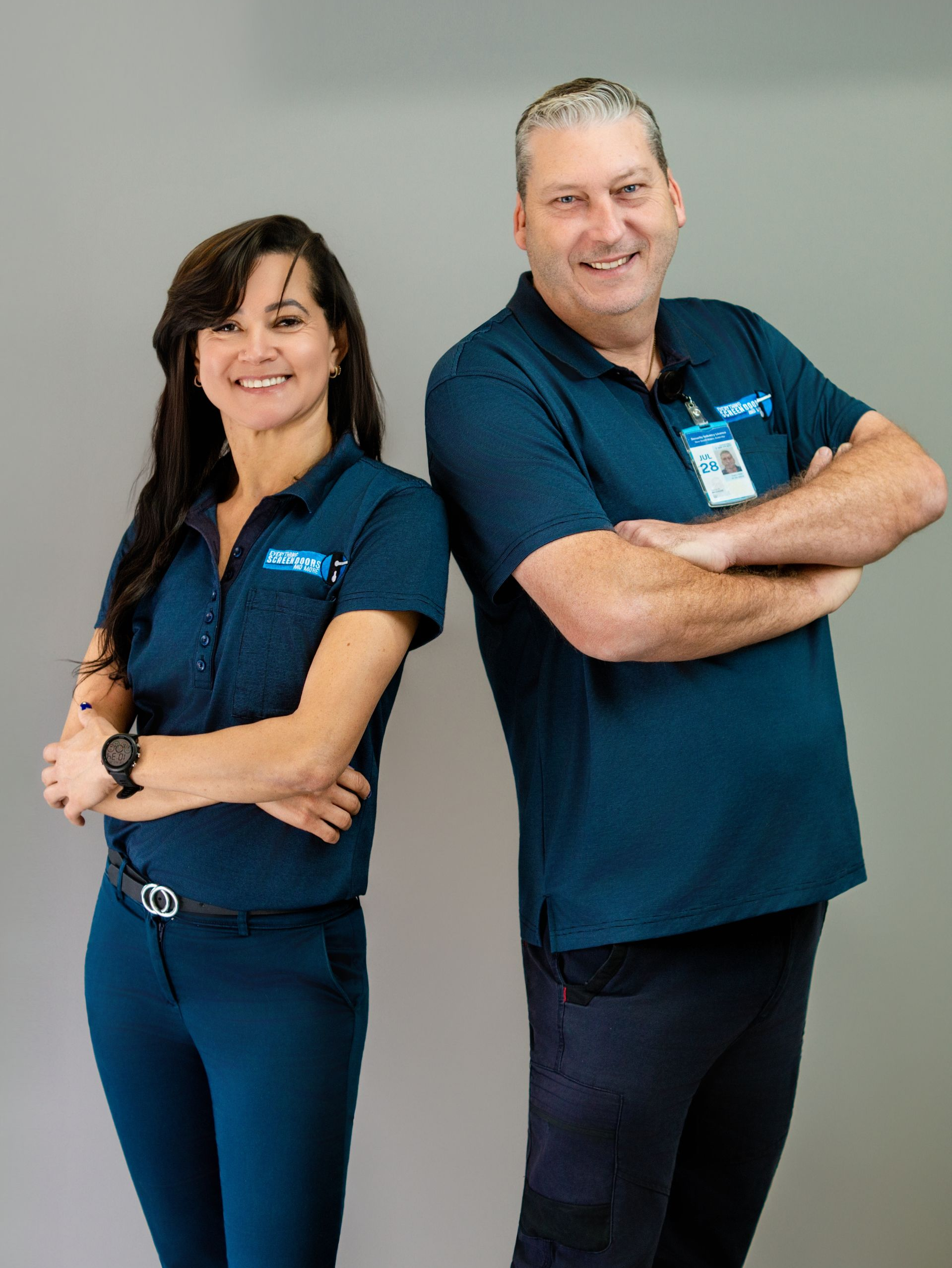 Woman and man in blue uniforms, arms crossed, smiling against a grey backdrop — Everything Screendoors & More In Wollongong, NSW