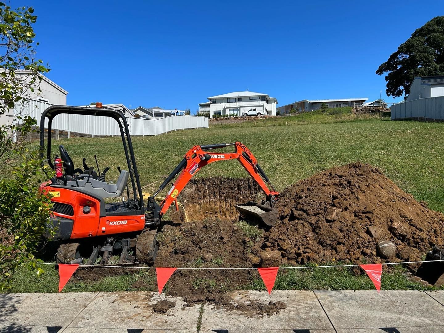 Orange excavator digging in a grassy area, pile of dirt, buildings in background, sunny day — JLH Plumbing, Gas & Roofing in South Nowra, NSW