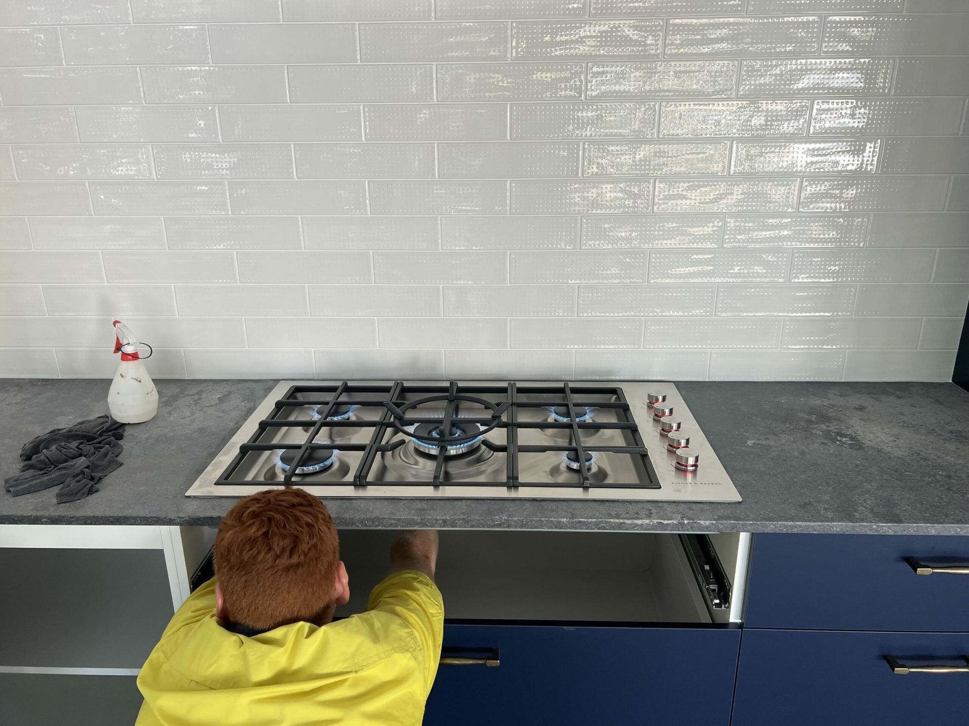A person installs a gas cooktop in a kitchen with blue cabinets and white brick-like tiles — JLH Plumbing, Gas & Roofing in South Nowra, NSW