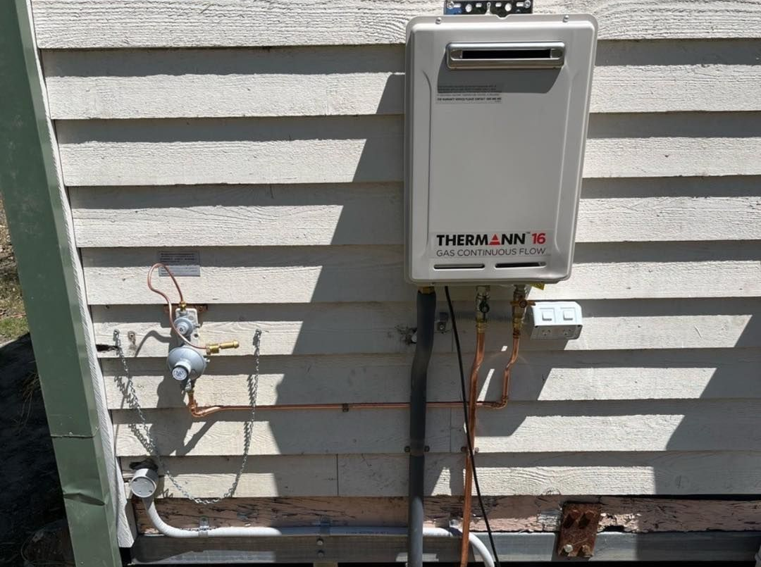 Tankless water heater mounted on a weathered, horizontal-slat siding, with associated pipes and gas meter — JLH Plumbing, Gas & Roofing in South Nowra, NSW