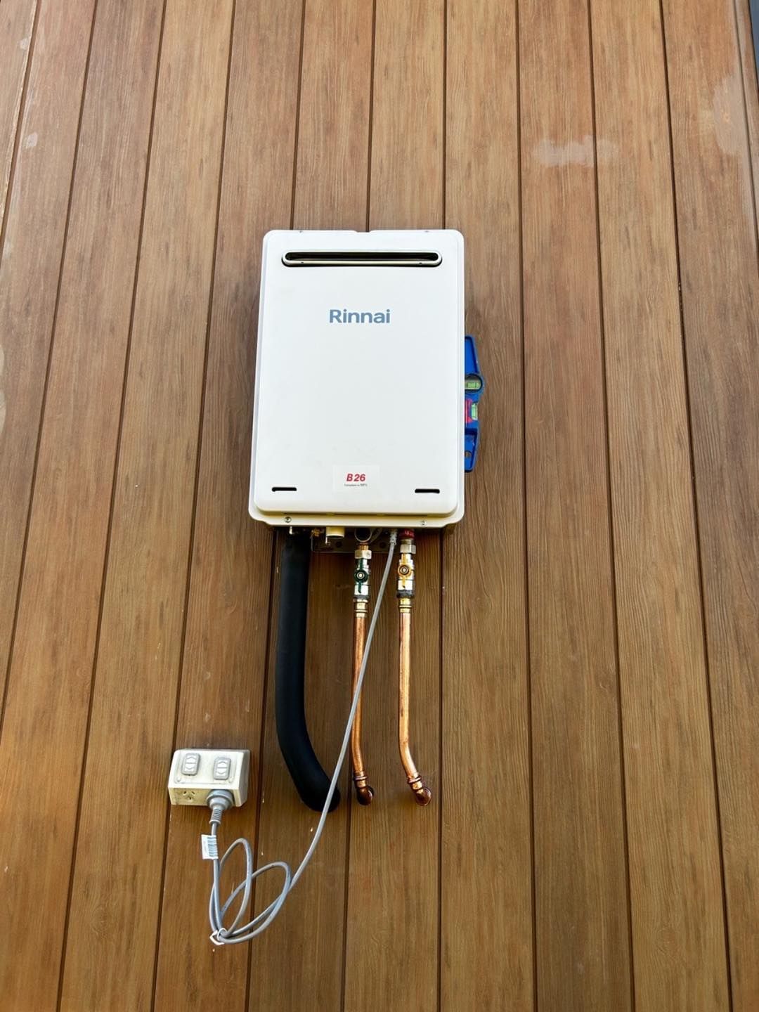 Rinnai tankless water heater mounted on wood siding with water and gas pipes, and electrical outlet — JLH Plumbing, Gas & Roofing in South Nowra, NSW