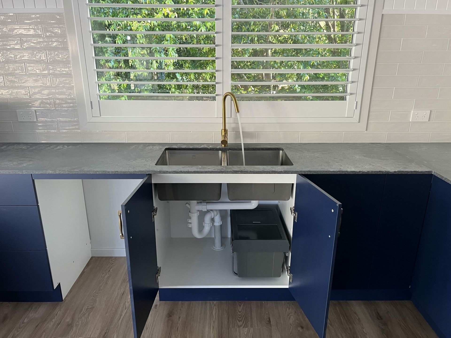 Open cabinet under a kitchen sink with plumbing and a trash can. Blue cabinets, grey countertop, gold tap — JLH Plumbing, Gas & Roofing in South Nowra, NSW