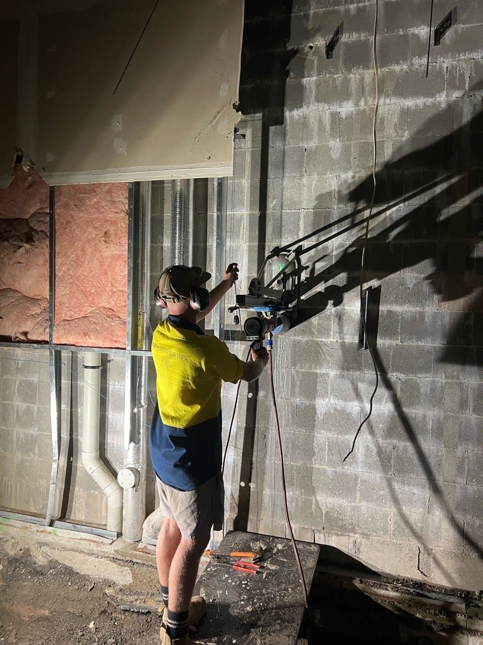 Man drilling into a cinder block wall, wearing a hard hat and safety gear, inside a building — JLH Plumbing, Gas & Roofing in South Nowra, NSW