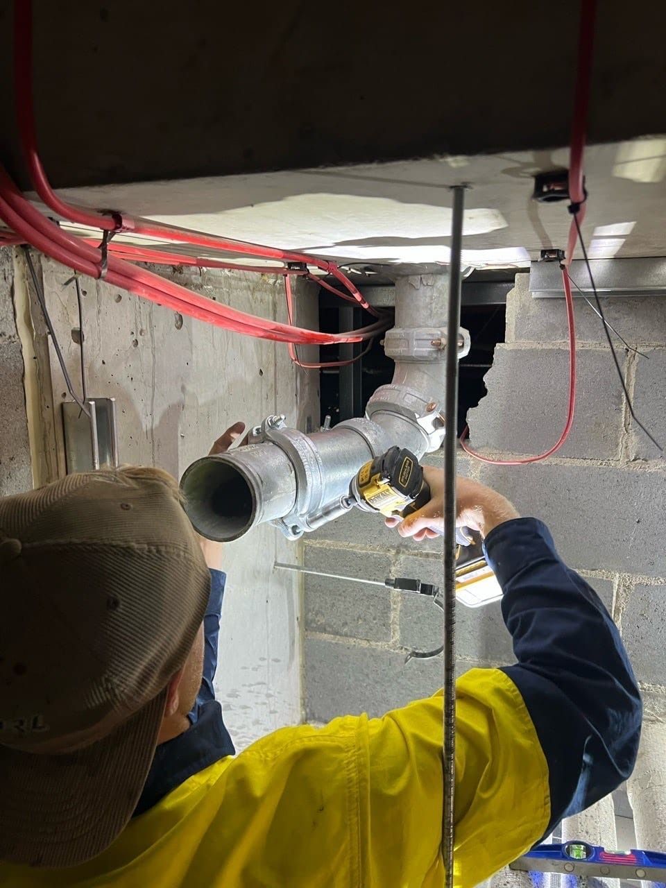 A person in yellow and blue workwear installs ductwork with a drill near red wires — JLH Plumbing, Gas & Roofing in South Nowra, NSW