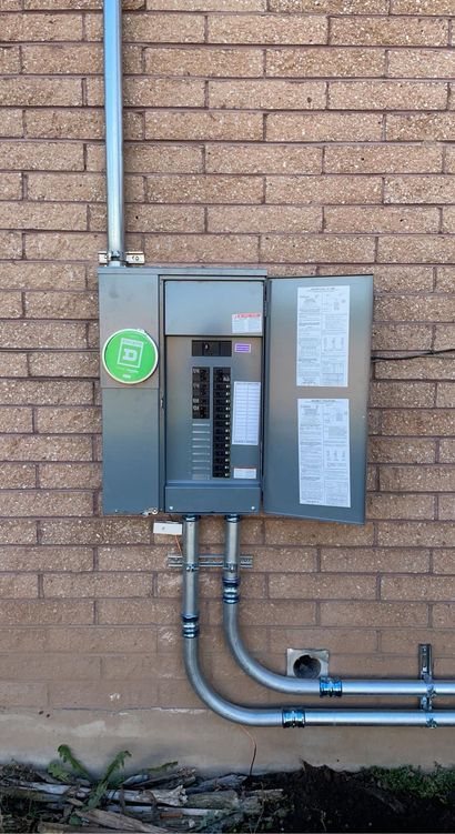 An electrical service panel mounted on a brick wall, with conduits extending from the top and bottom.