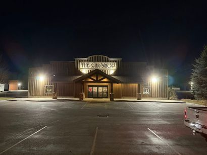 A dark, outdoor parking lot view of a rustic, wood-paneled building at night with a bright, illuminated sign reading.