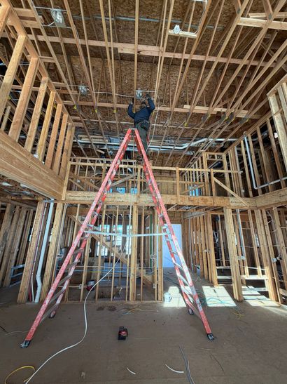 A person stands on a tall ladder working on electrical wiring in the exposed wood framing of a house under construction.