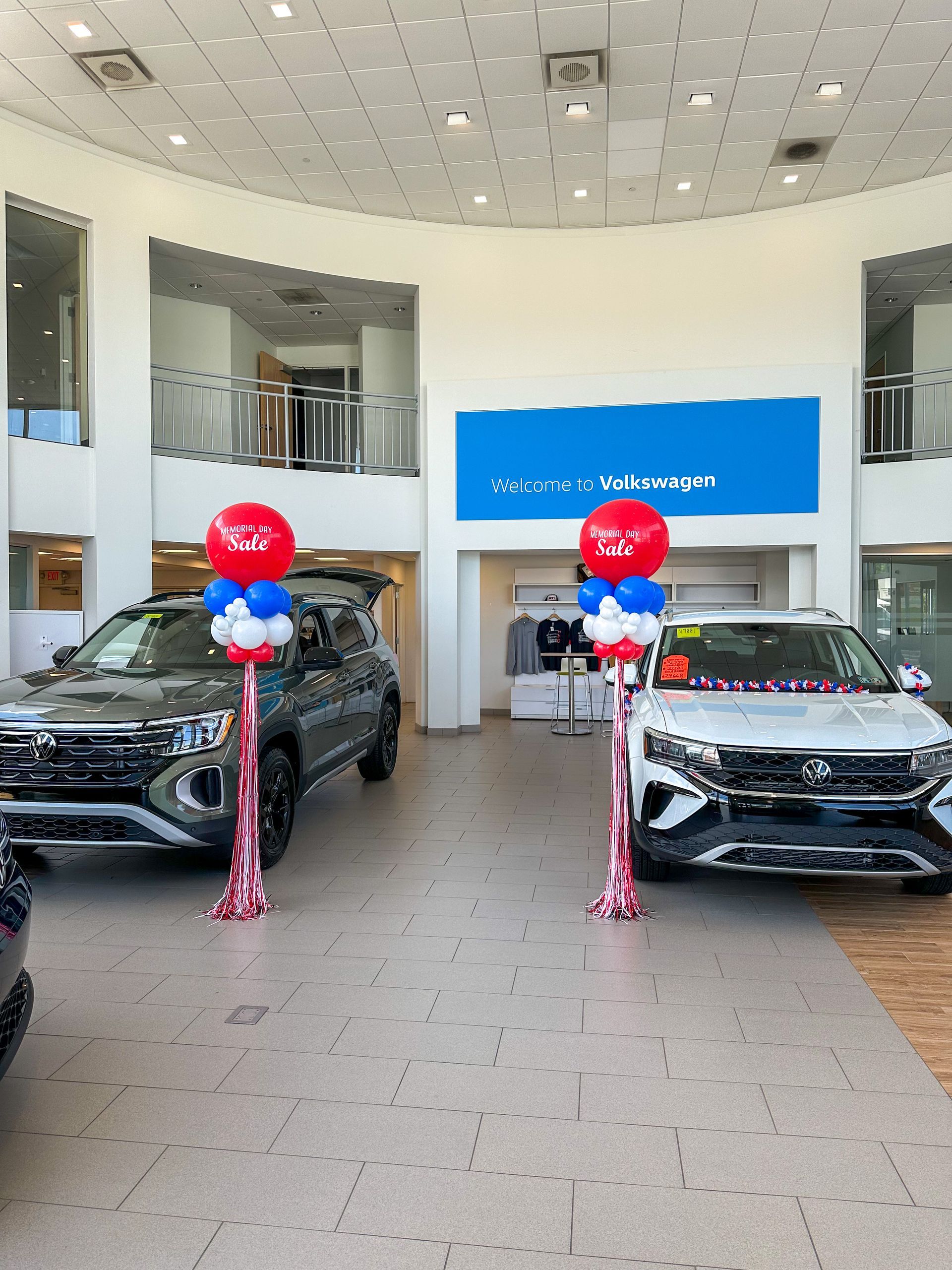 Two cars are parked in a showroom with red , white and blue balloons.