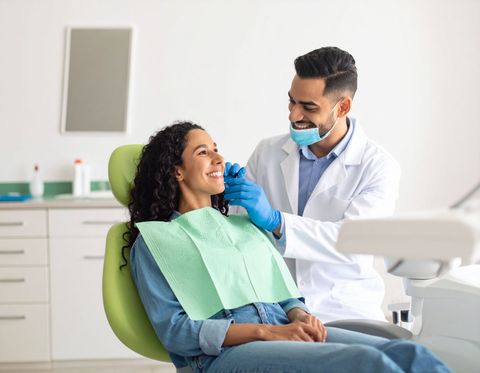 Dentist examining a smiling patient in a dental chair; white coat, blue gloves, modern office.