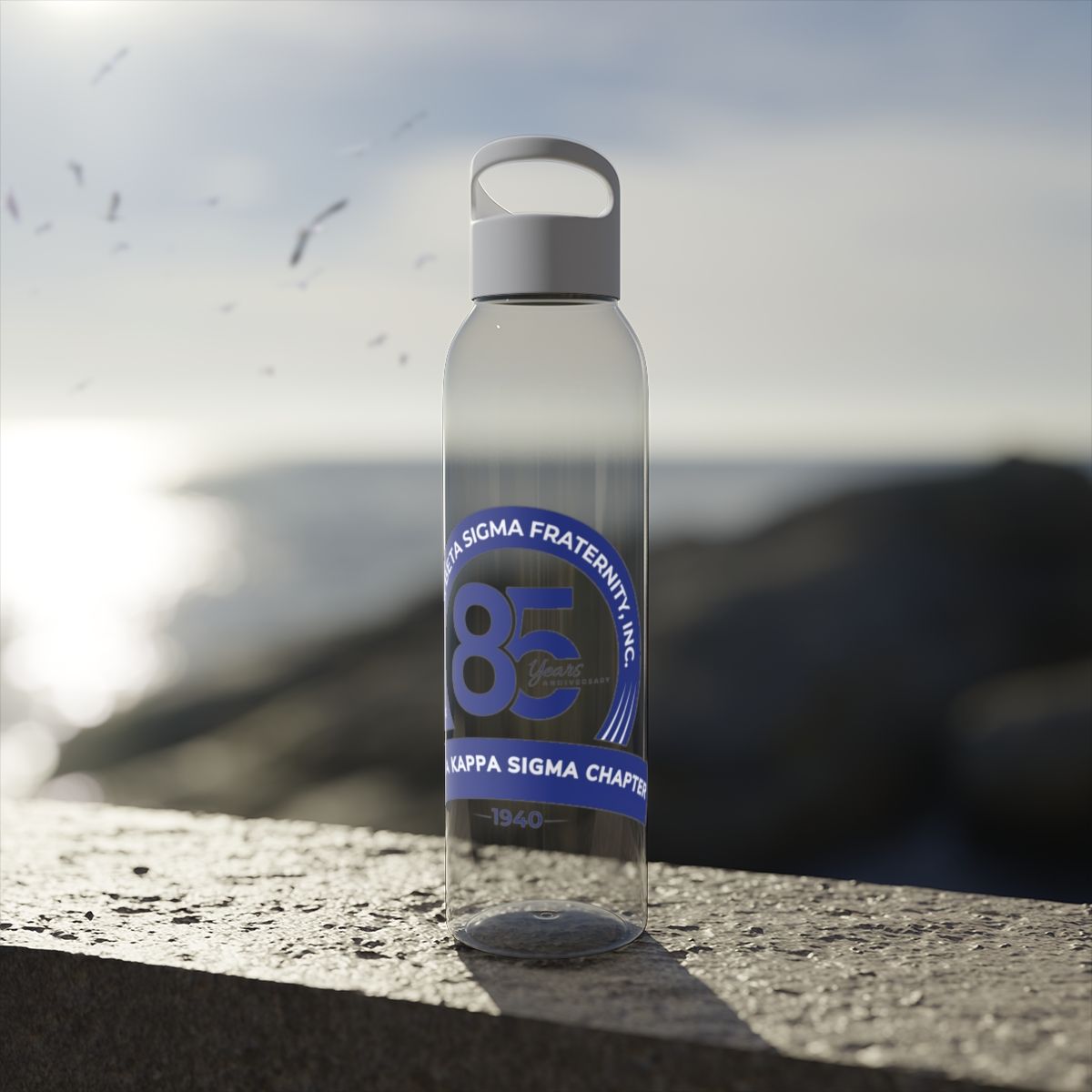 Clear water bottle with blue fraternity logo on a stone wall, beach background.