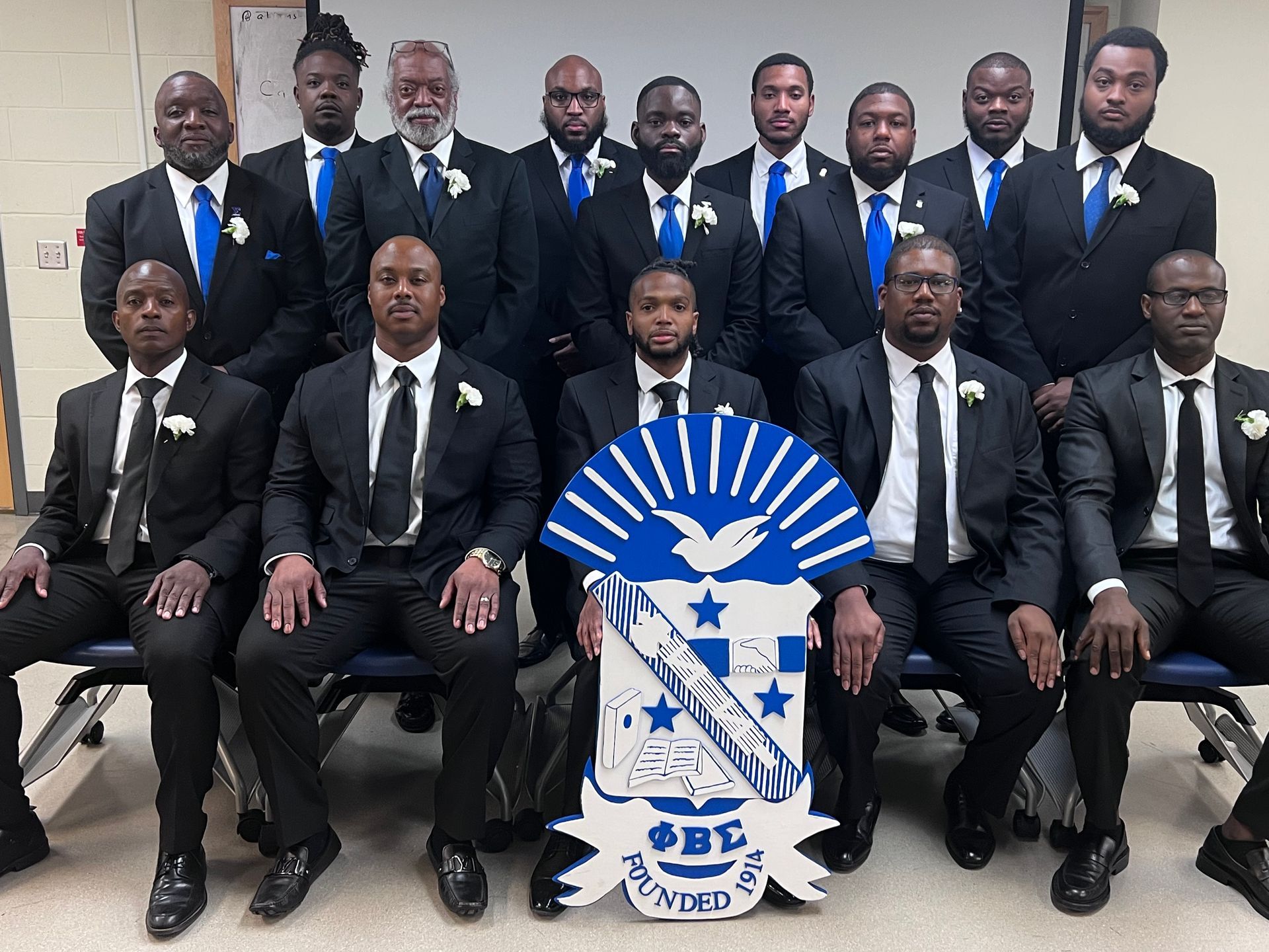 Group of men in black suits with blue ties, posing with a blue and white crest, indoors.