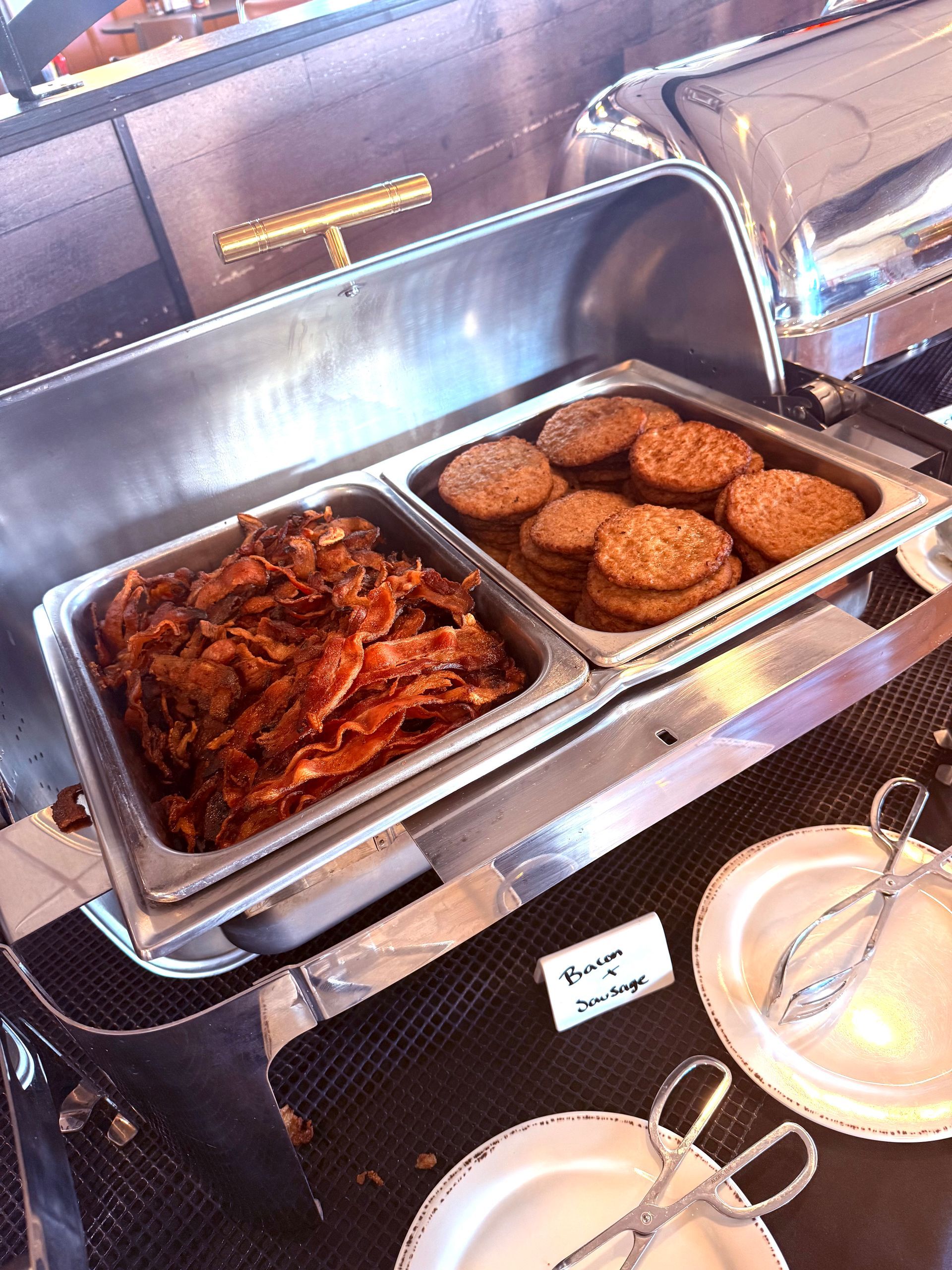 A hotel breakfast buffet warmer containing crispy bacon and round sausage patties, with serving tongs and plates.