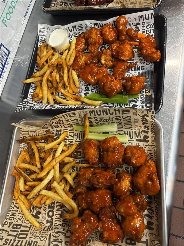 Two trays of boneless buffalo wings with french fries and celery sticks, served on branded paper liners.