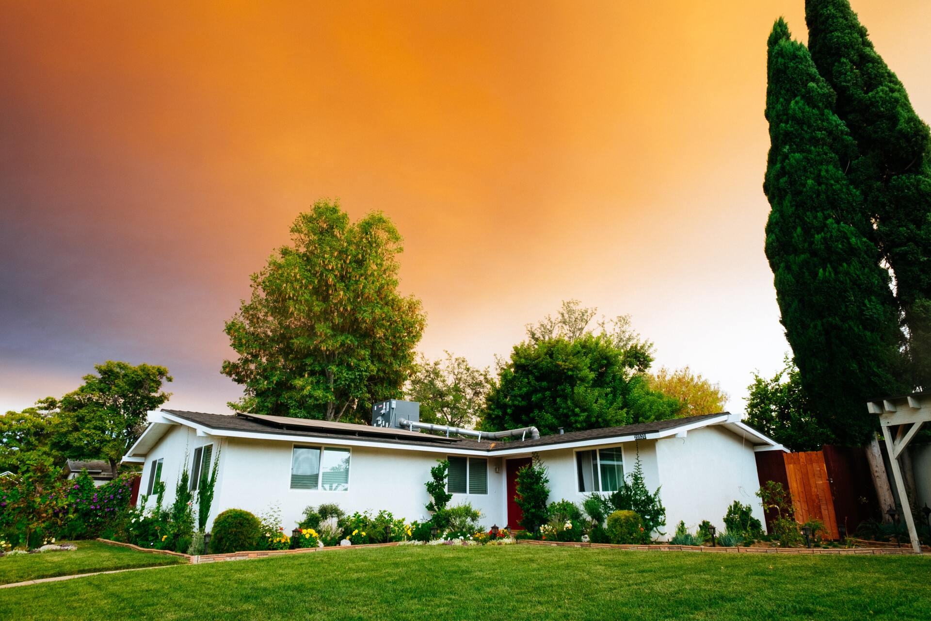 Sunset casts a warm orange glow over a charming house with a lush green lawn and majestic trees.