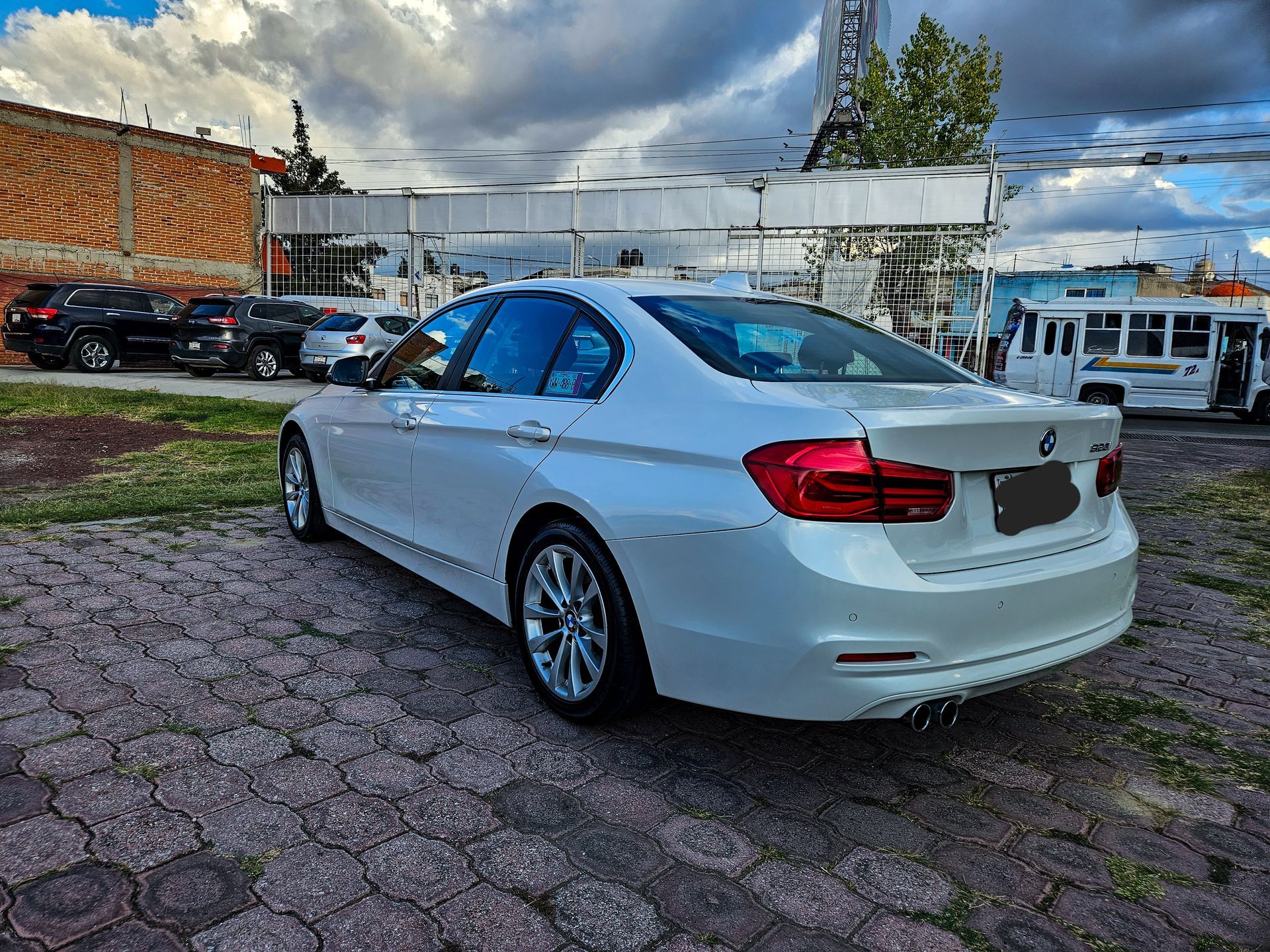 BMW sedán blanco estacionado sobre un suelo de ladrillo, con otros vehículos y un edificio al fondo. Cielo nublado.