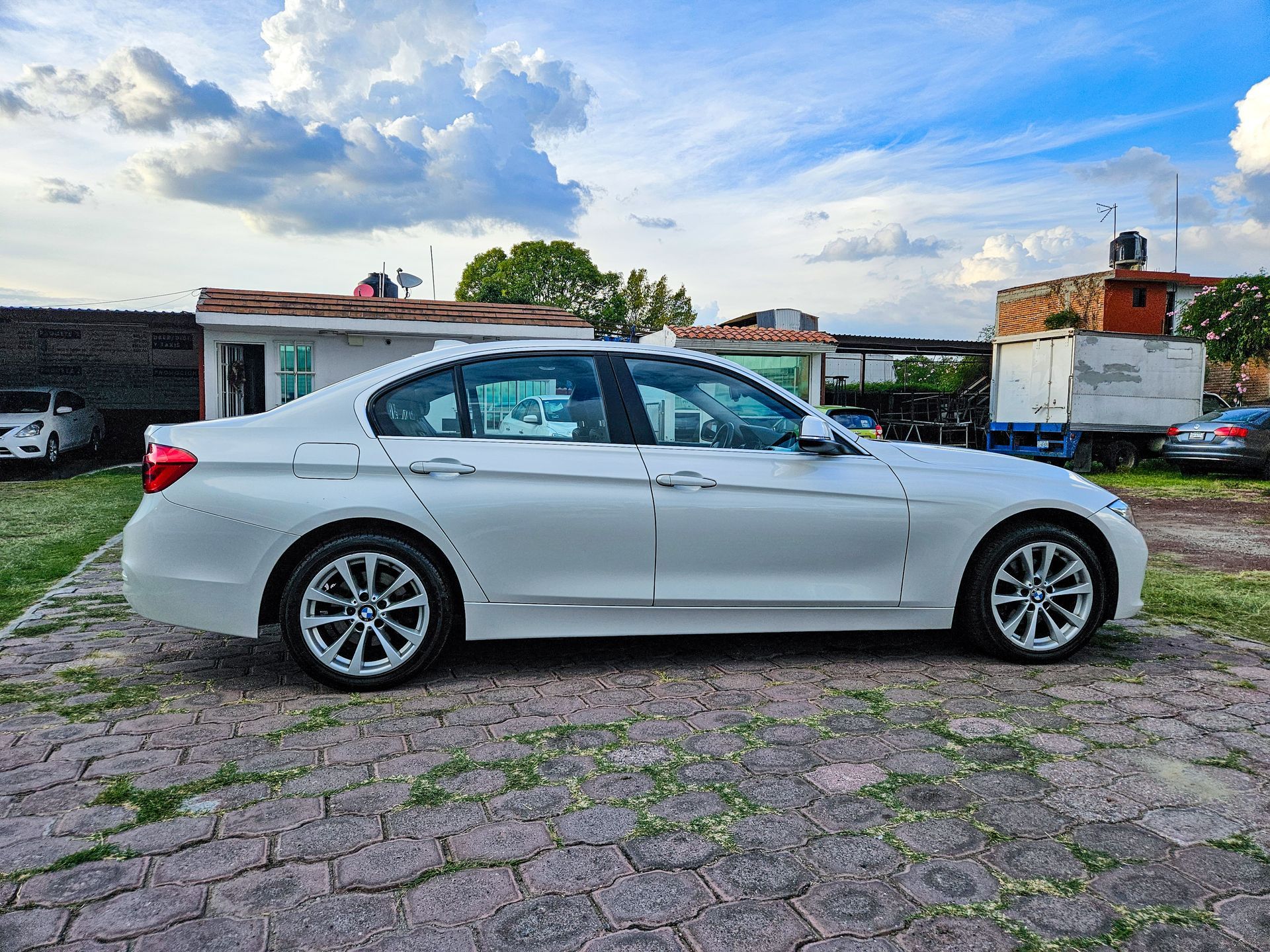 Sedán BMW blanco estacionado sobre una superficie de ladrillo con edificios y cielo al fondo.
