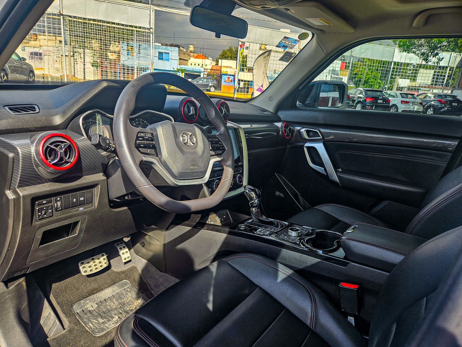 Vista interior de un coche negro con volante, tablero y asientos. Se aprecian detalles en rojo y molduras de fibra de carbono.