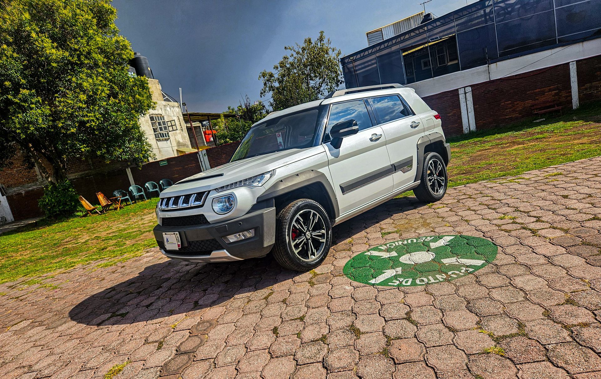 SUV blanca estacionada en un camino adoquinado, vista frontal con llantas negras. Césped verde, árboles y edificios al fondo.