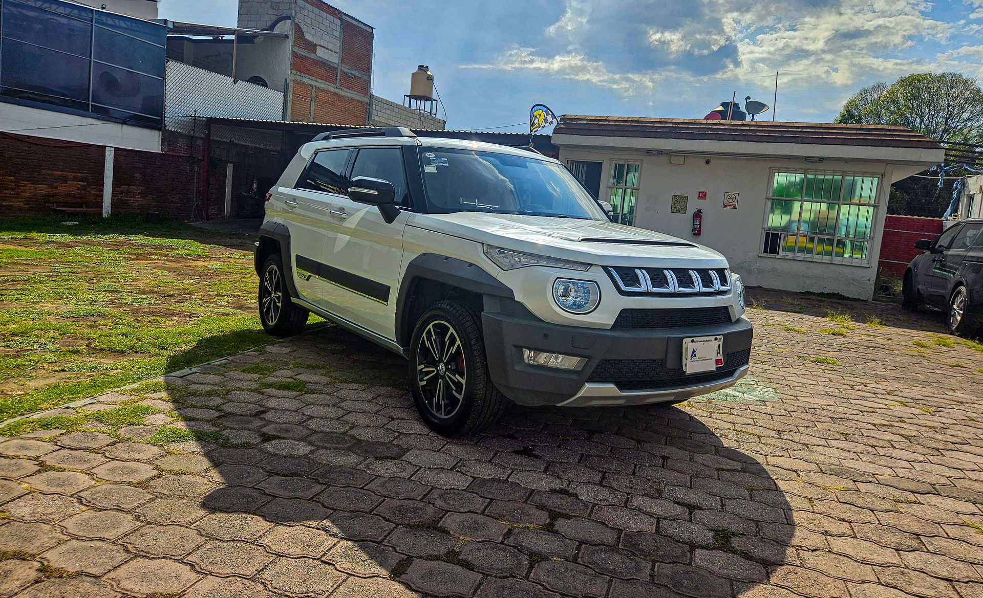 SUV blanco estacionado en un área pavimentada con ladrillos frente a un pequeño edificio.