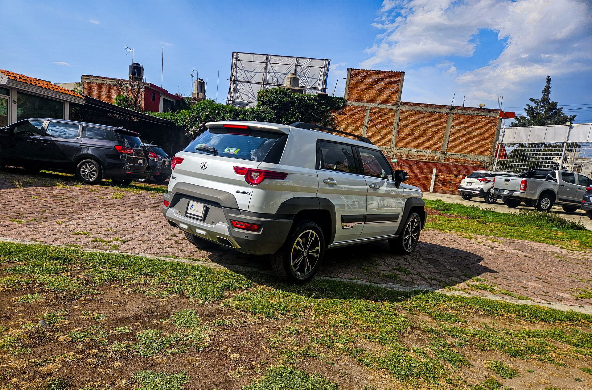 SUV plateado estacionado en un lote de grava junto a otros vehículos, con edificios y un cielo nublado al fondo.