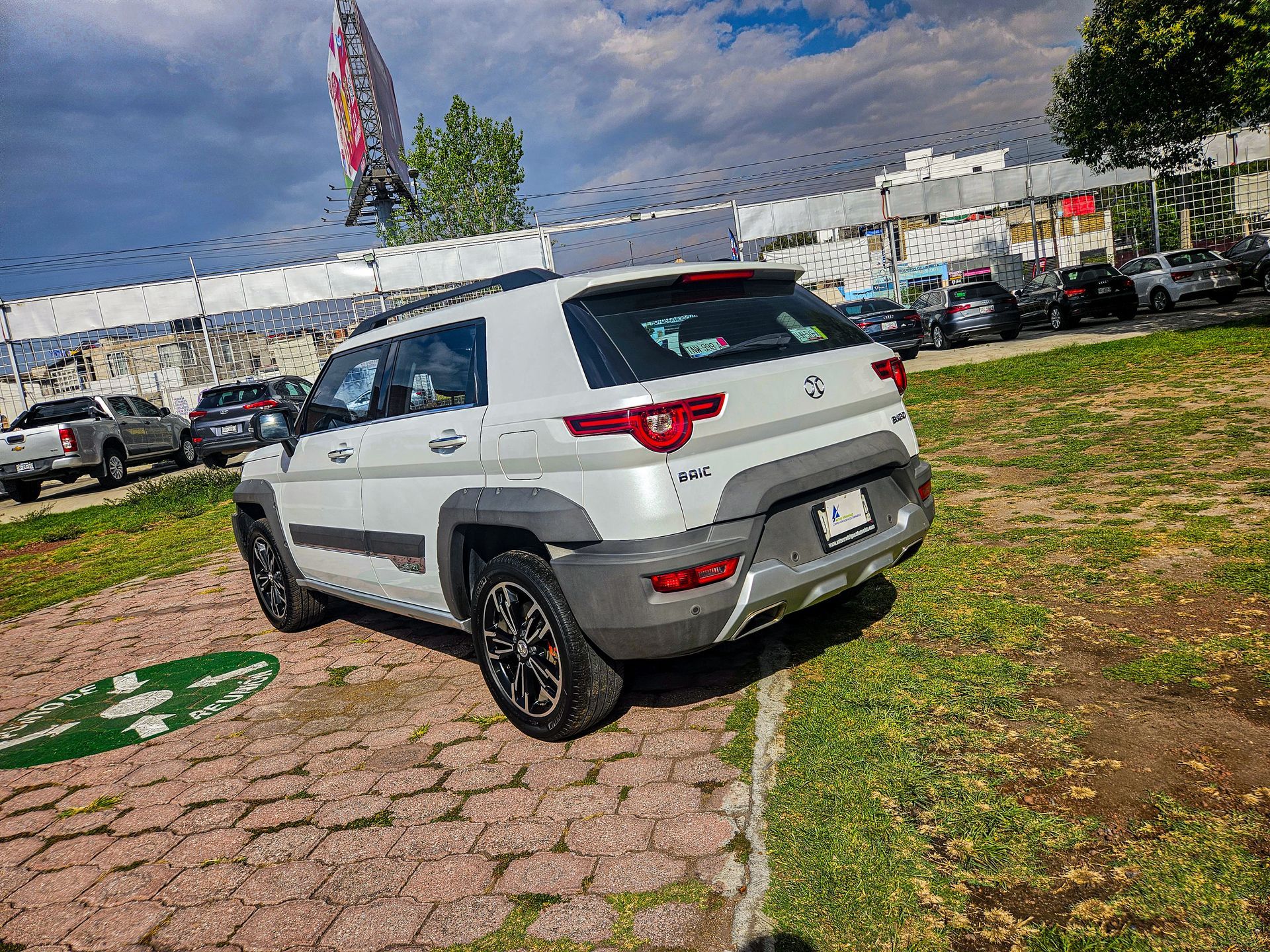 Una camioneta blanca estacionada sobre ladrillos y césped cerca de un concesionario. Detalles en negro y cielo nublado.