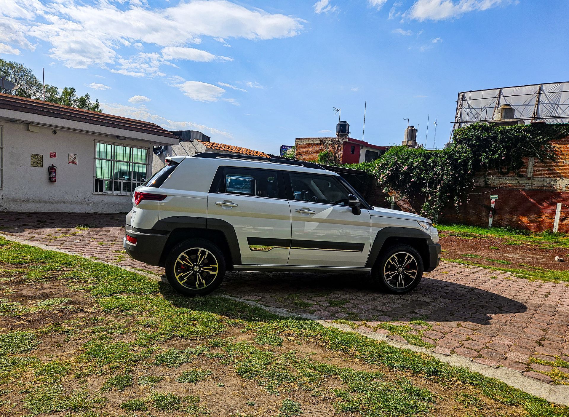 Una camioneta blanca estacionada sobre el césped junto a un camino de ladrillos. Edificio con vegetación y cielo azul al fondo.