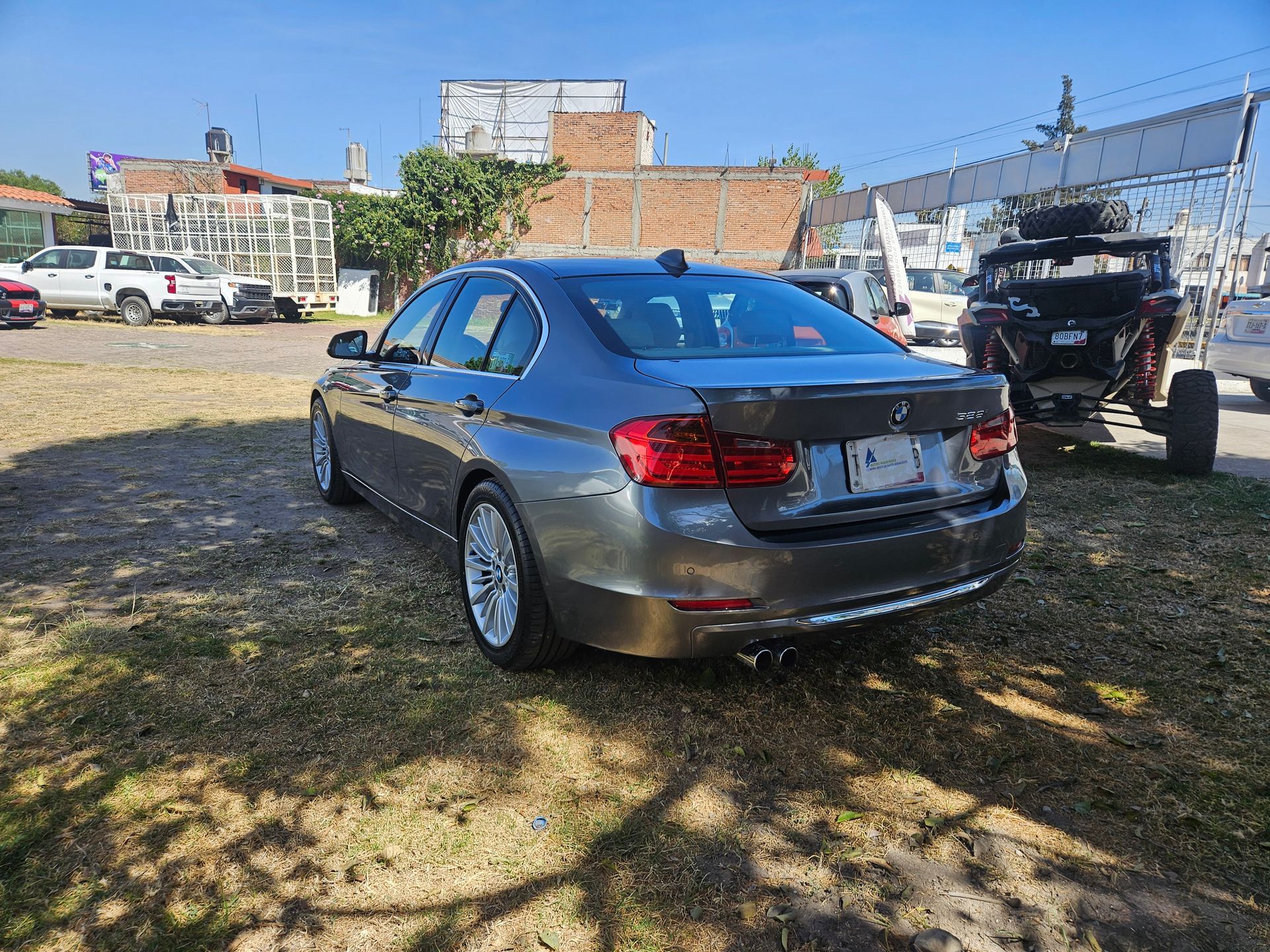 Sedán BMW gris estacionado en el césped con otros vehículos y edificios al fondo.
