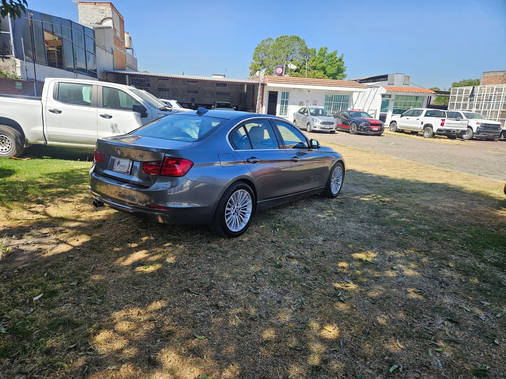 Sedán gris estacionado en una zona verde, rodeado de otros vehículos y edificios bajo un cielo soleado.