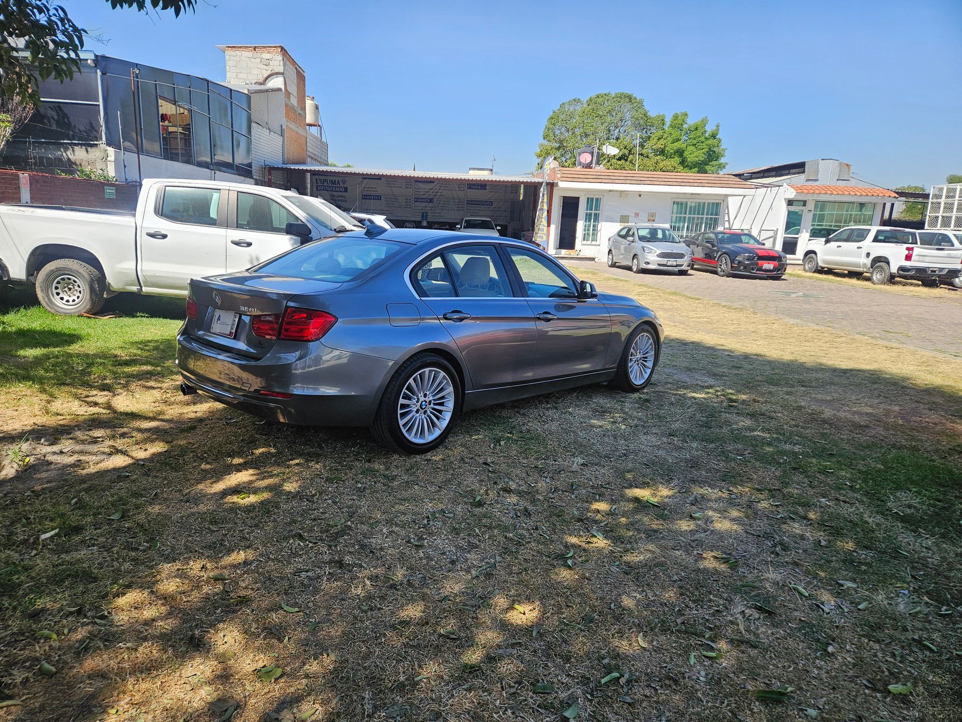Sedán gris estacionado en una zona verde, con otros vehículos y edificios al fondo. Día soleado.