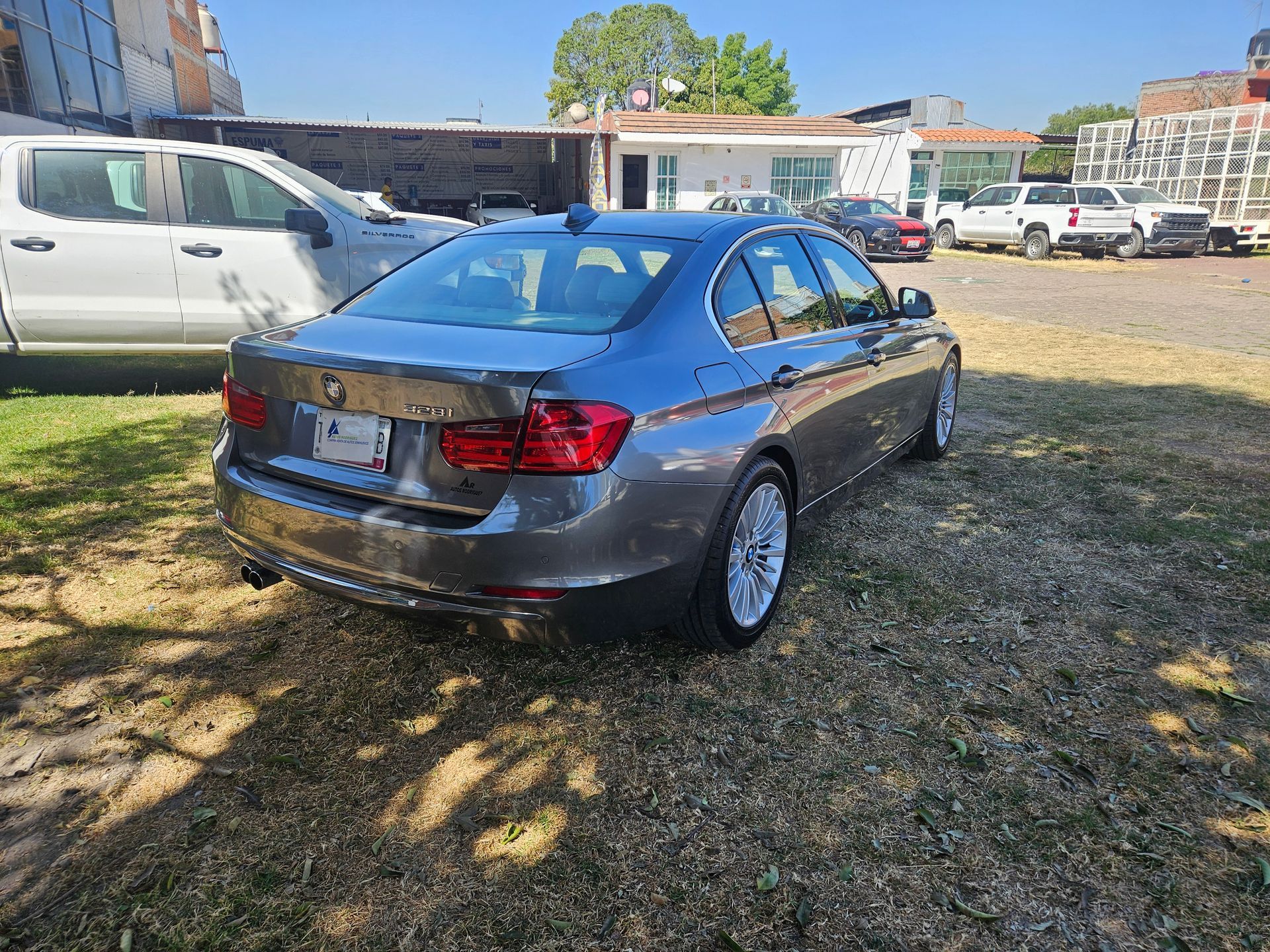 Un sedán BMW gris estacionado en un terreno con césped, con otros vehículos y edificios al fondo.