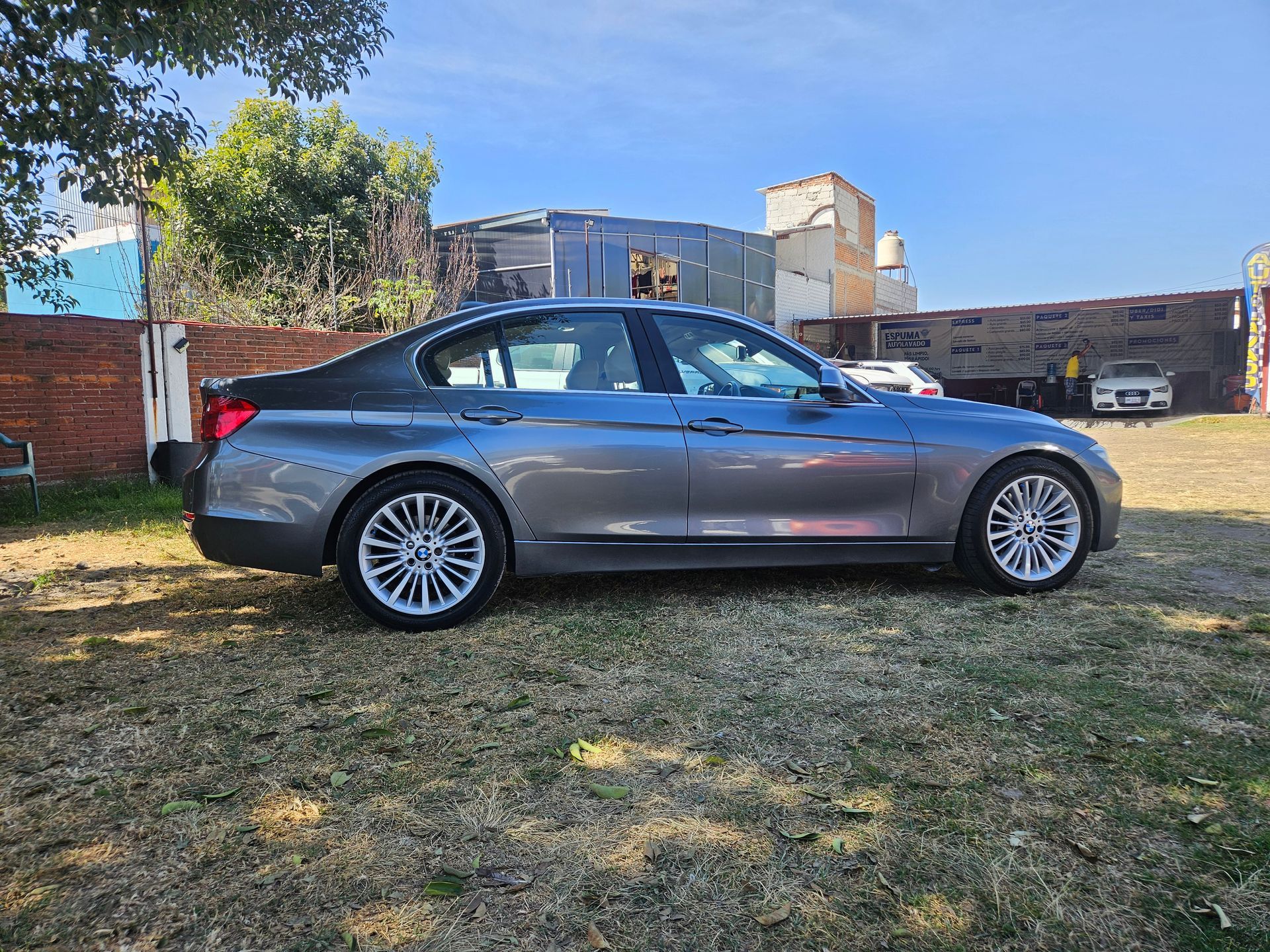 Un sedán BMW gris estacionado sobre el césped con una pared de ladrillos y edificios industriales al fondo.