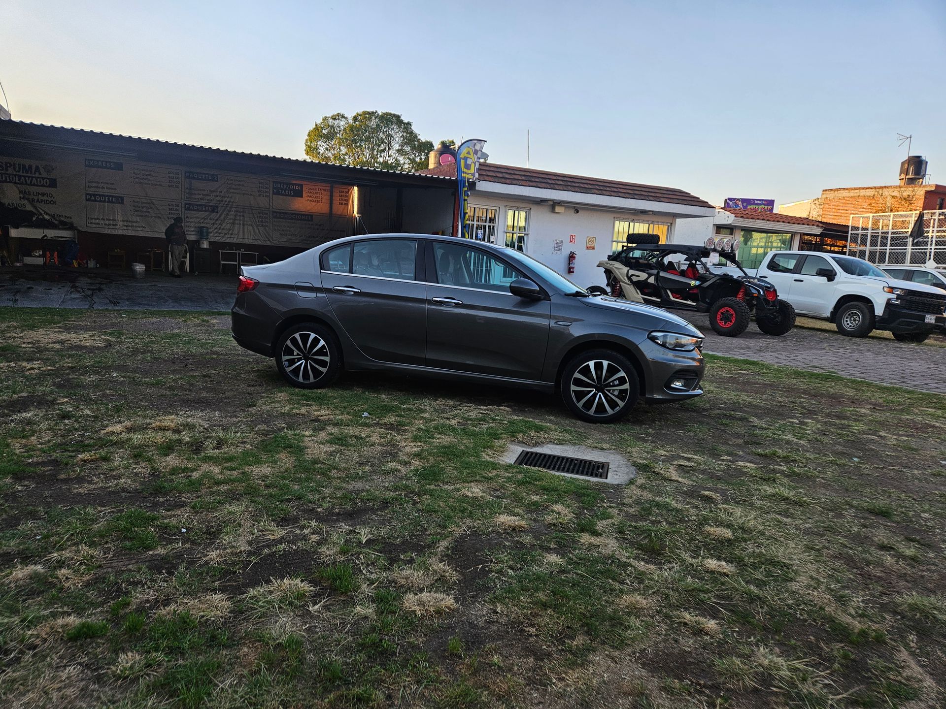 Sedán gris estacionado en un área con césped cerca de edificios y otros vehículos.