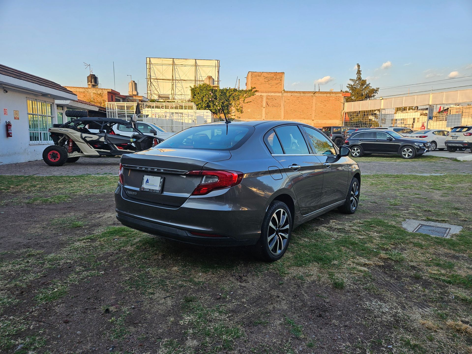 Automóvil sedán gris estacionado sobre el césped, con otros vehículos y edificios al fondo.