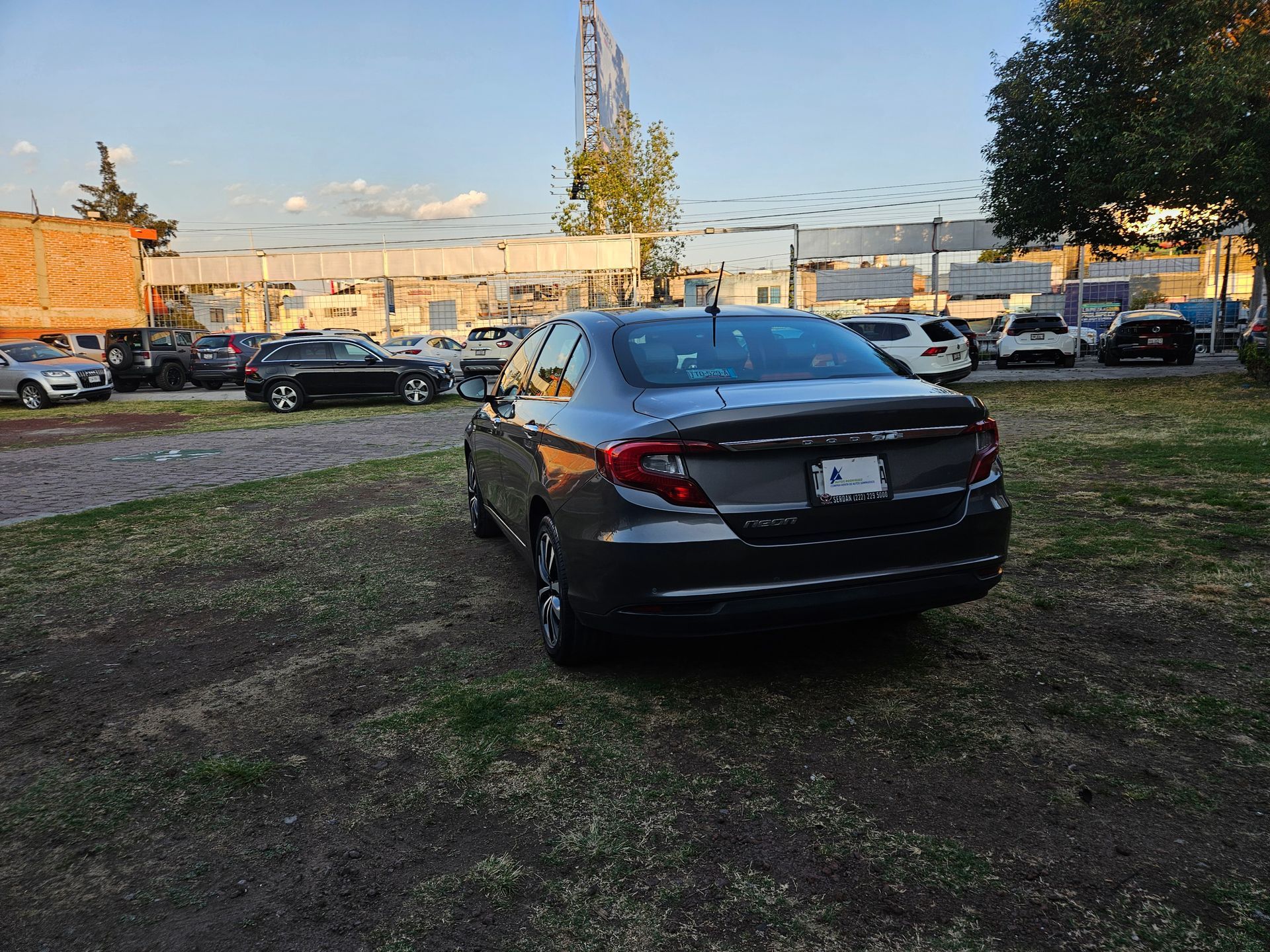 Sedán oscuro estacionado en un estacionamiento con césped; otros autos y un edificio al fondo.