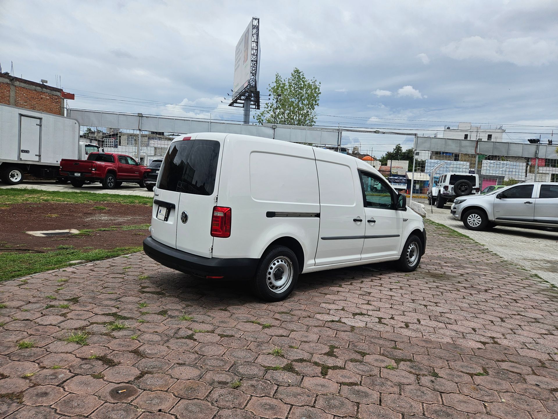 Furgoneta de carga blanca estacionada sobre ladrillo. Cielo nublado, otros vehículos y un cartel al fondo.
