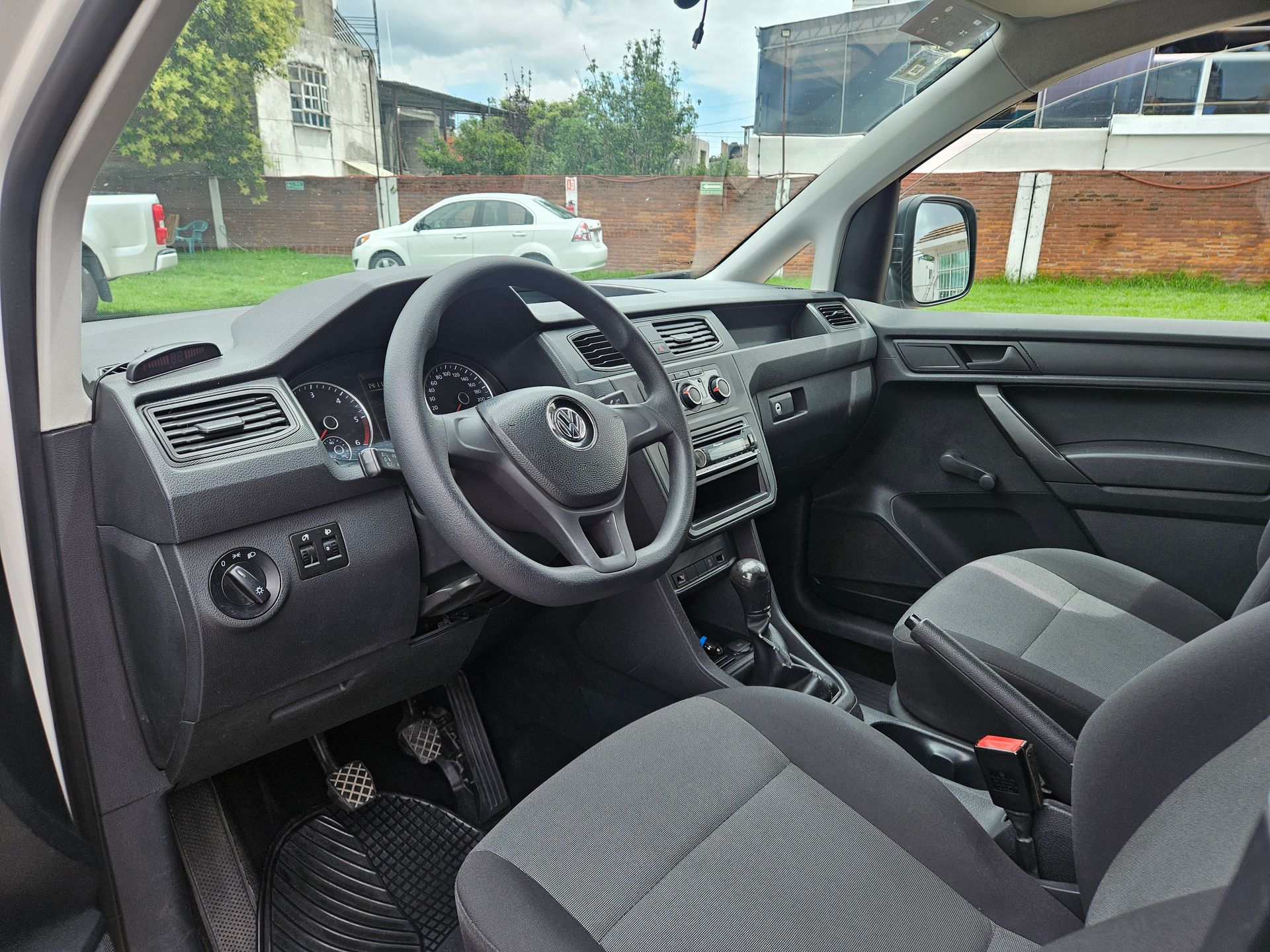 Vista interior de un Volkswagen Caddy. Tablero, volante y asientos en negro y gris. Vista exterior desde la ventana.