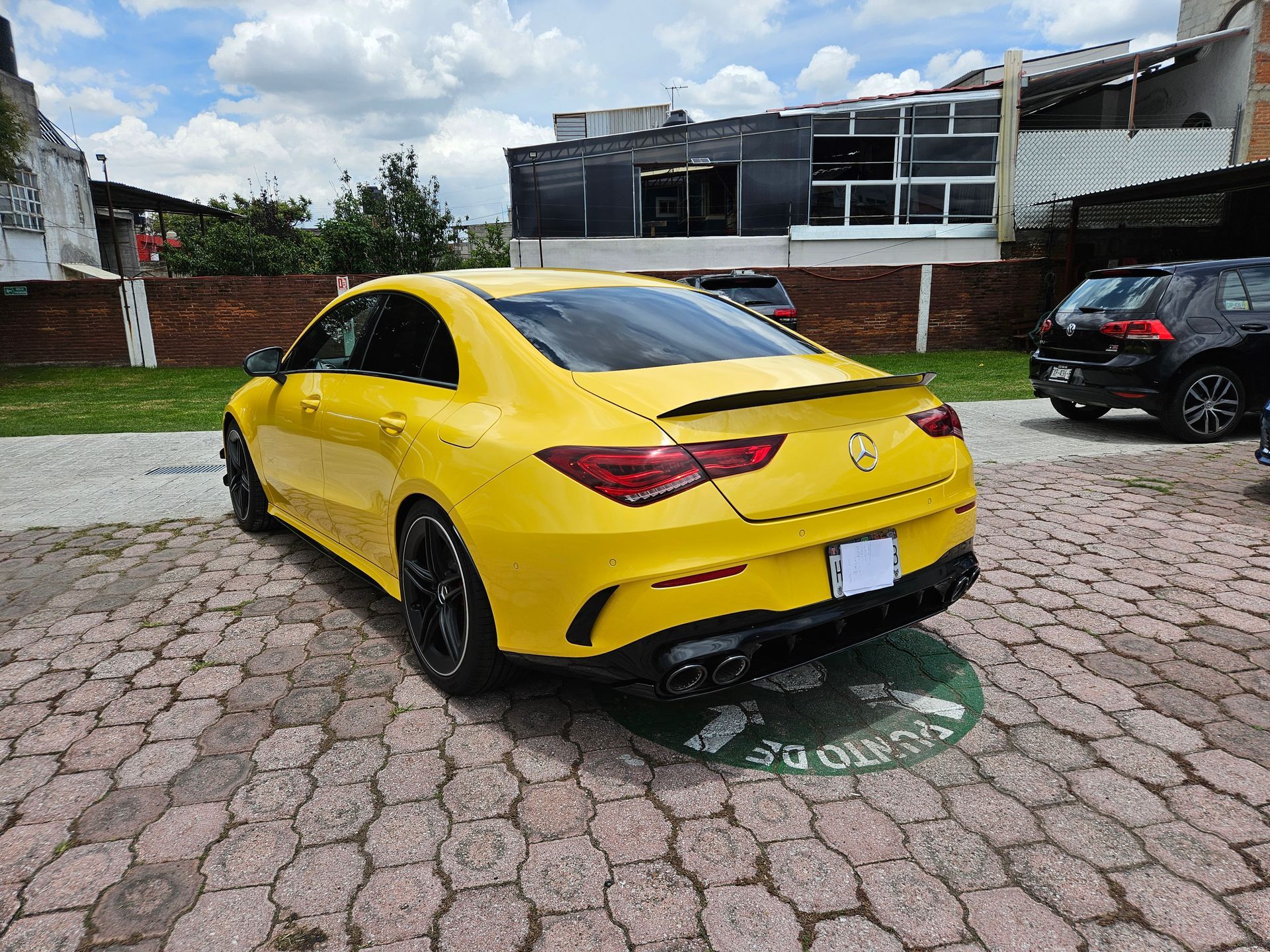 Mercedes-Benz amarillo estacionado en una zona pavimentada con ladrillos, cerca de un edificio y otros vehículos en un día soleado.