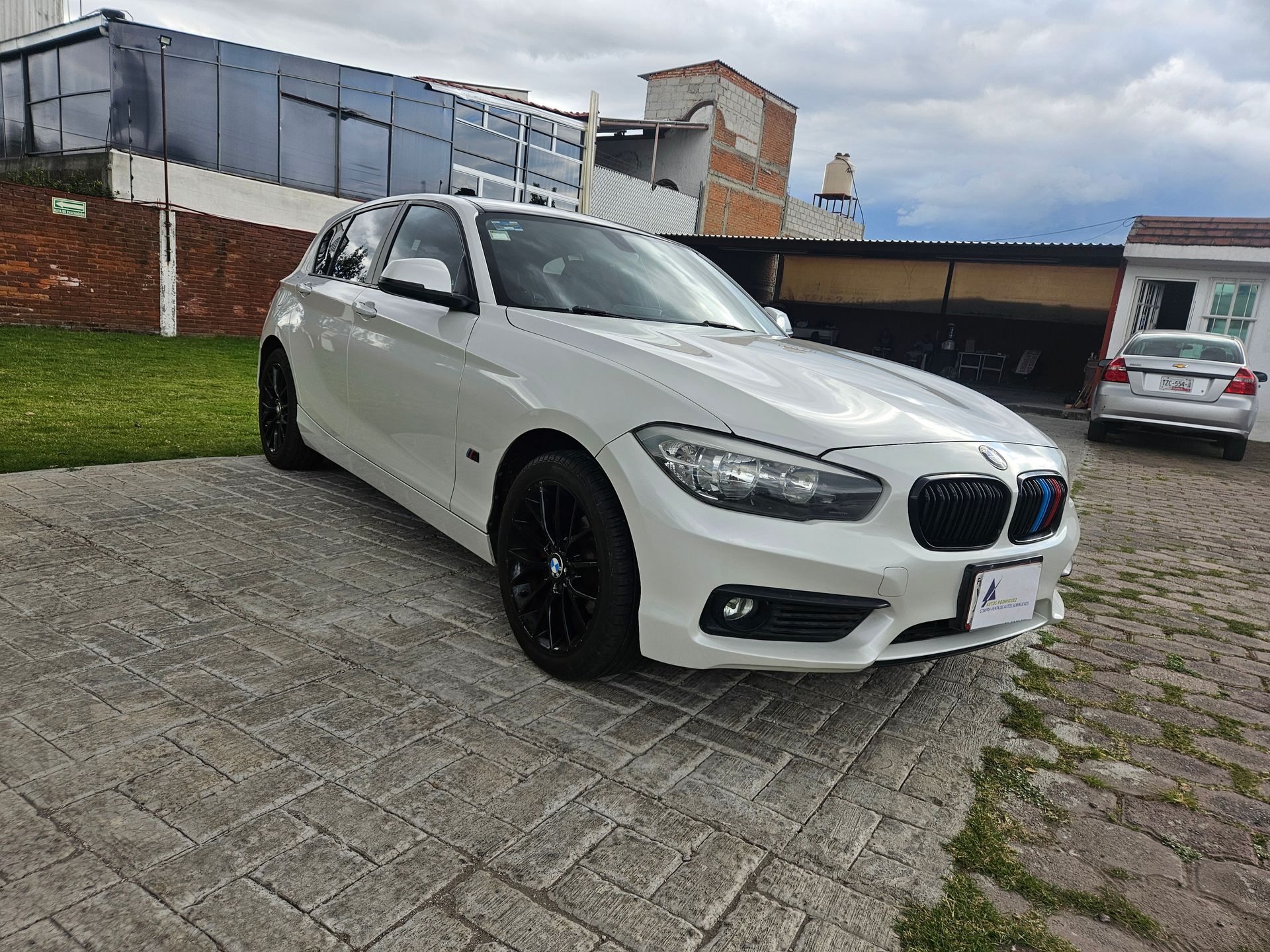 BMW hatchback blanco estacionado en una entrada pavimentada; cielo nublado.