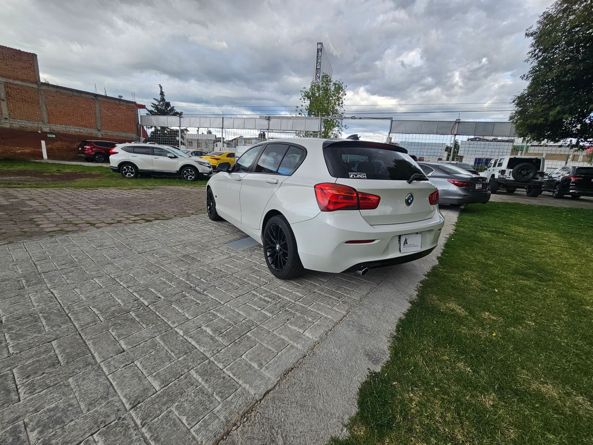 BMW hatchback blanco estacionado sobre una superficie de ladrillo junto a la hierba, con fondo de cielo nublado.