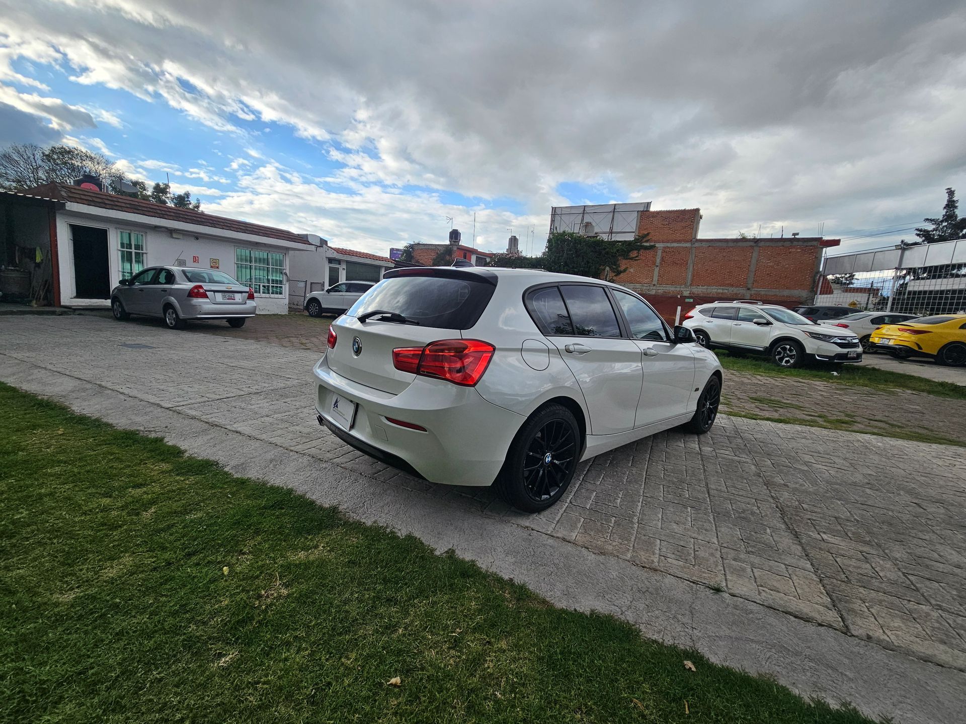Un BMW blanco hatchback estacionado en una entrada pavimentada; otros vehículos y edificios al fondo.